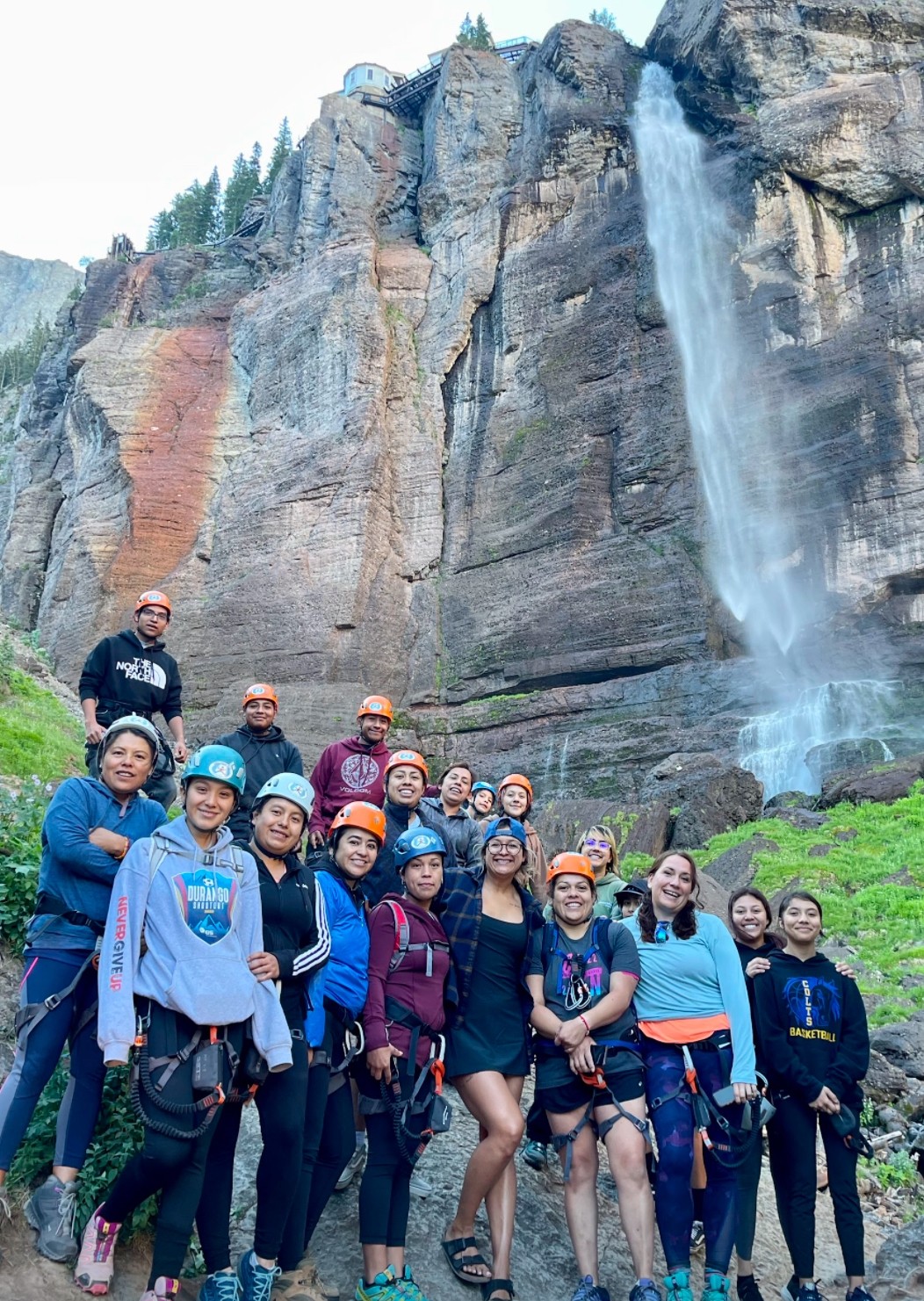 A group of Spanish-speaking community members standing in front of a waterfall on a Via Ferrata